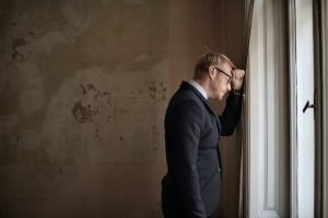 man in suit leaning head on wall next to window